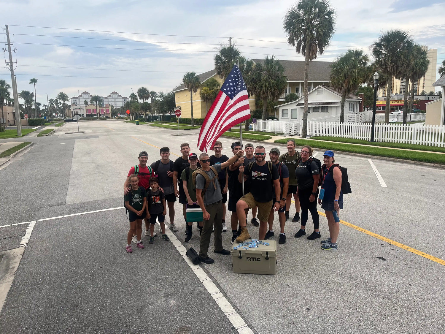Group of diverse GORUCK participants standing on a street holding an American flag with palm trees and houses in the background