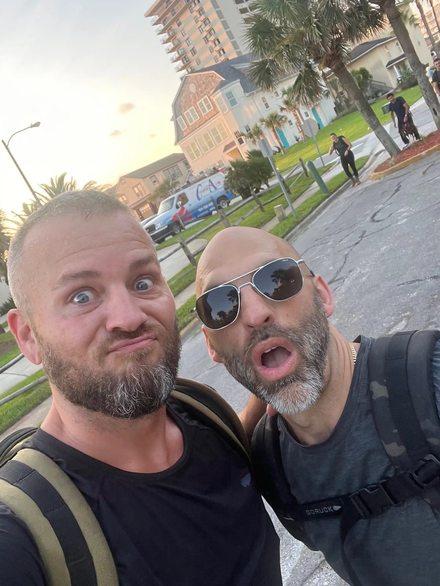 Two bearded men wearing backpacks, one with sunglasses, posing for a selfie outdoors near residential buildings at sunset