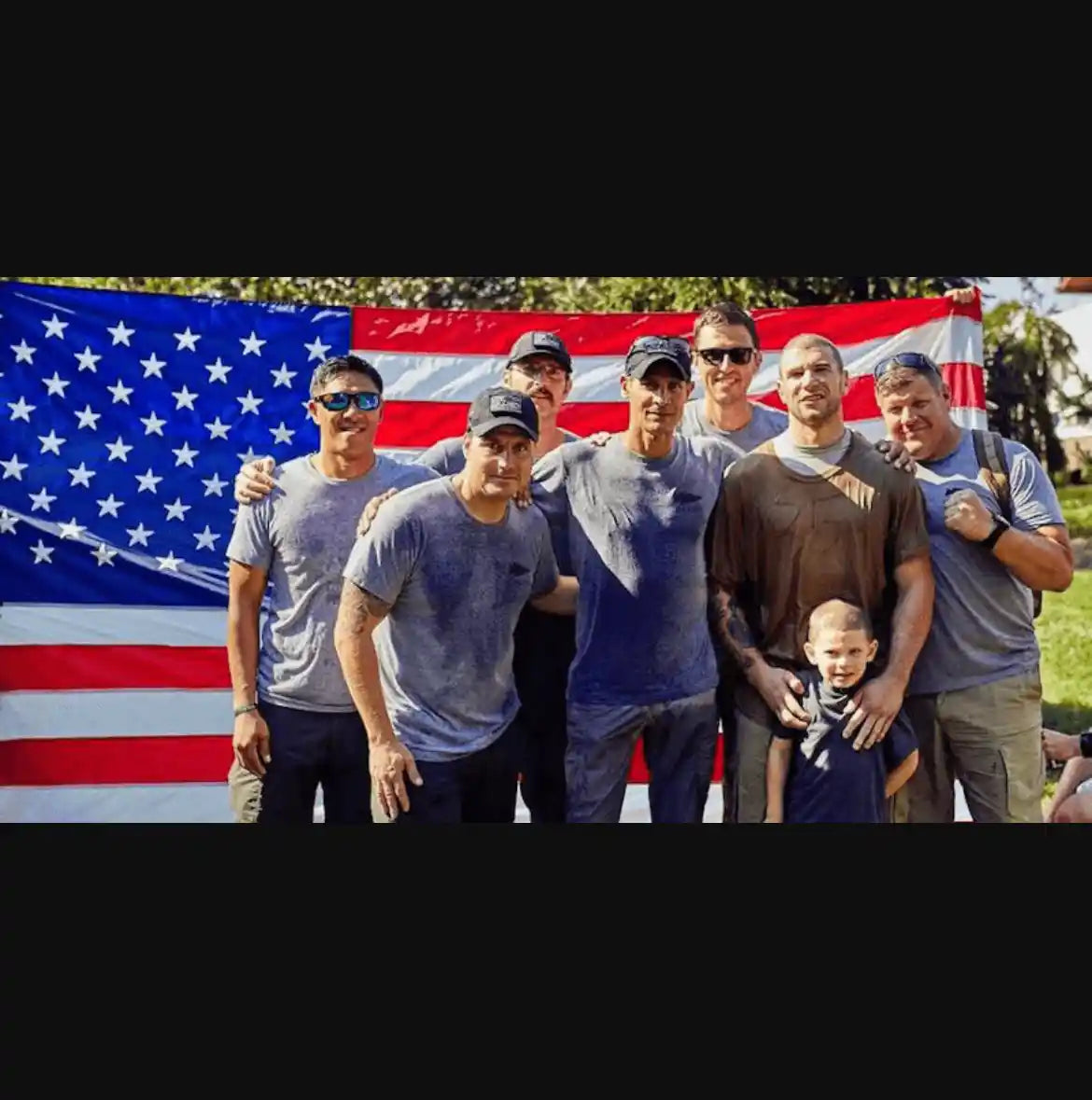 Group of men and a child in athletic gear posing in front of an American flag at a GORUCK event outdoors.