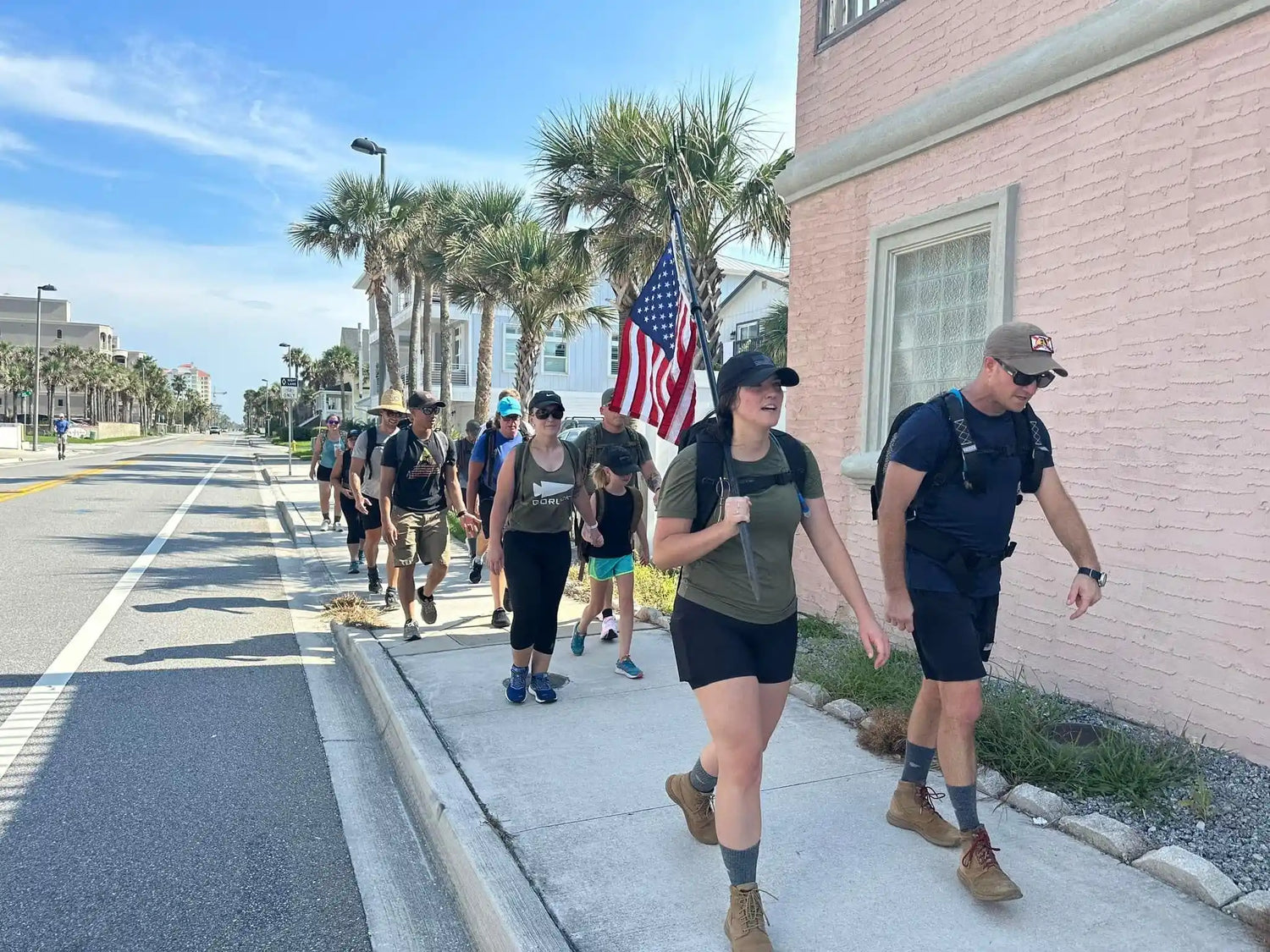 Group of people rucking in GORUCK gear, walking on city sidewalk with American flag, palm trees