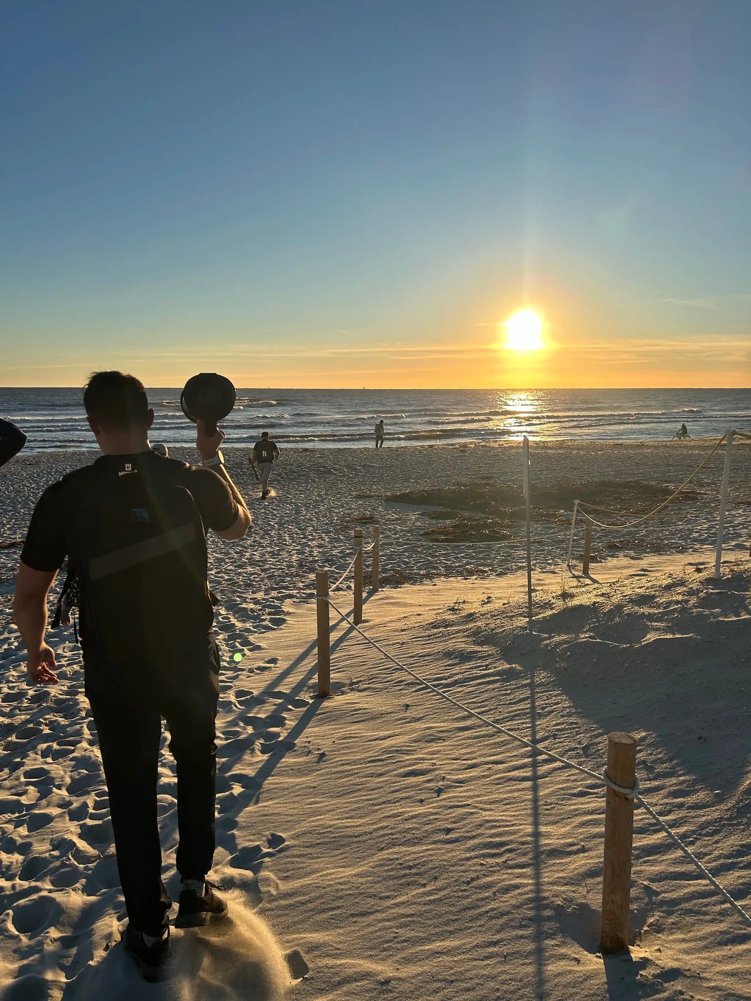 Person walking on sandy beach at sunset holding a hat with ocean and sun on horizon