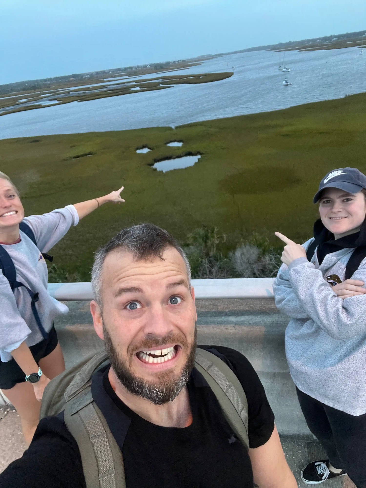 Three people with backpacks smiling on a bridge overlooking marshland and water, GORUCK rucking gear visible.