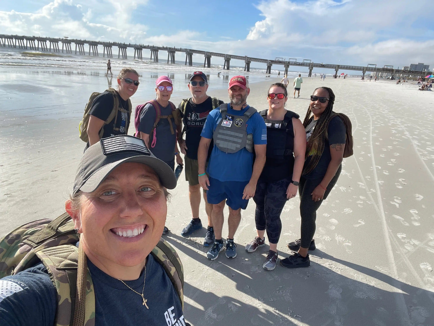 Group of GORUCK participants with backpacks at the beach near a pier under sunny skies