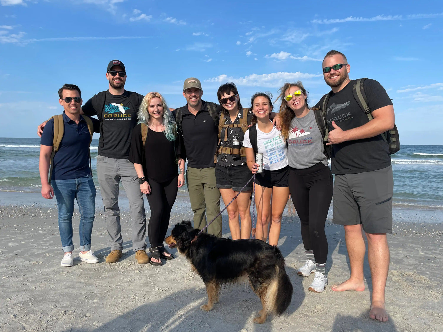 GORUCK team group smiling with backpacks and a dog on a sandy beach by the ocean