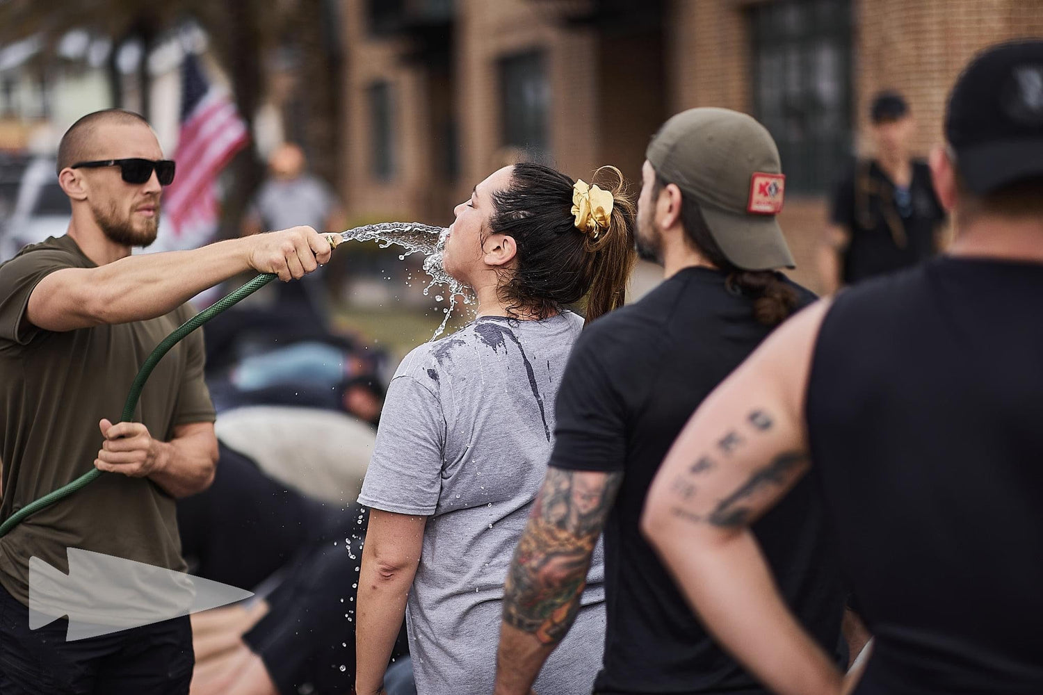 Outdoor GORUCK rucking training, woman drinks from hose, group in athletic gear.
