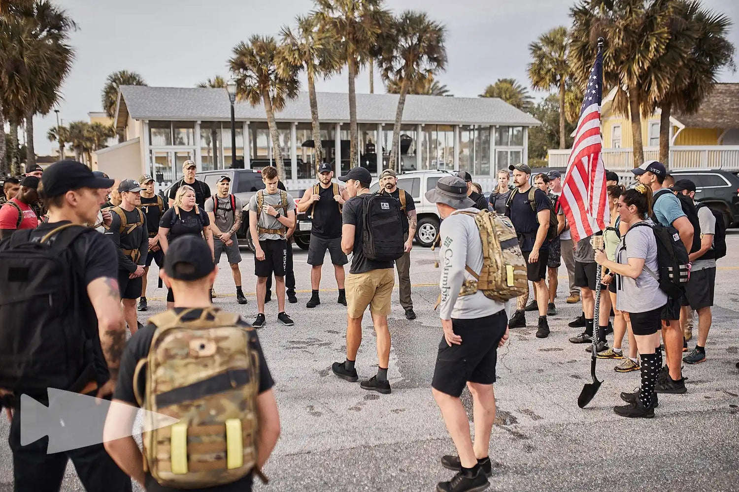 Group of people with backpacks in an outdoor urban area with palm trees, one holding an American flag