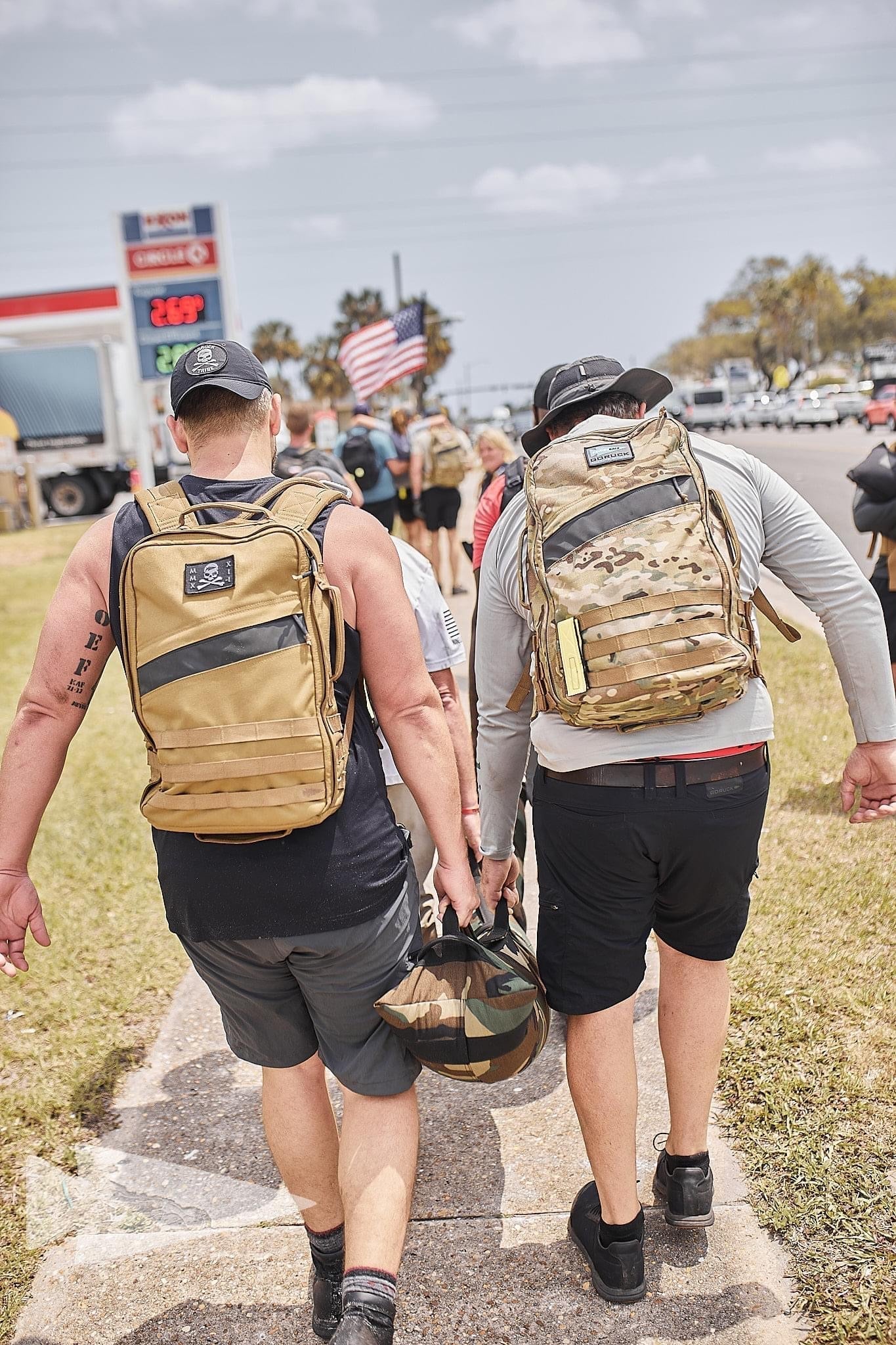 Group rucking with GORUCK backpacks on sidewalk, American flag visible, sunny day.