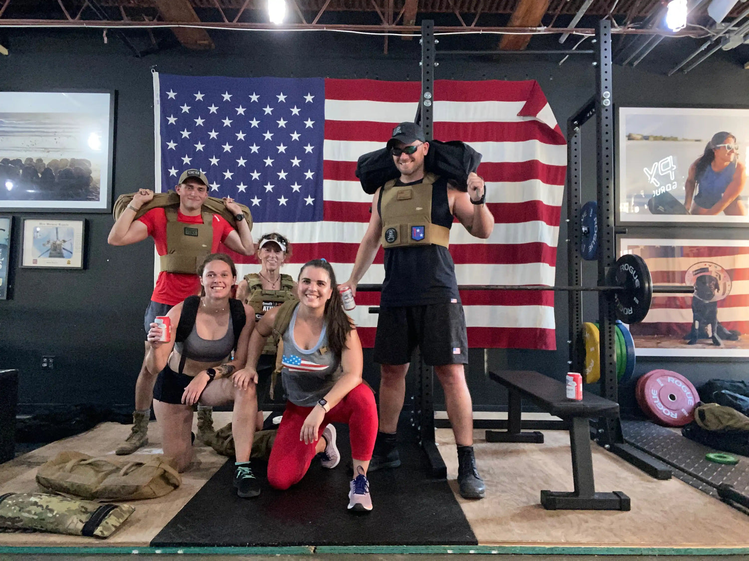 Group wearing GORUCK gear posing in gym with American flag background, fitness equipment visible.