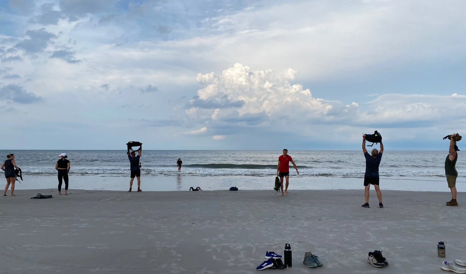 Group training with GORUCK gear on beach, people lifting rucksacks near ocean under cloudy sky.