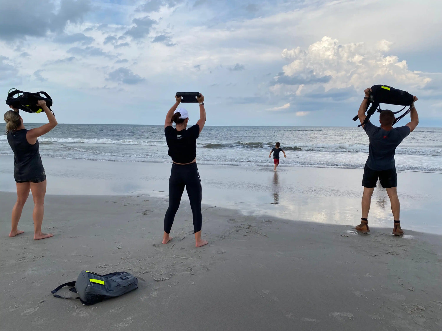 Three people rucking with GORUCK gear on a beach, ocean waves and clouds in background.