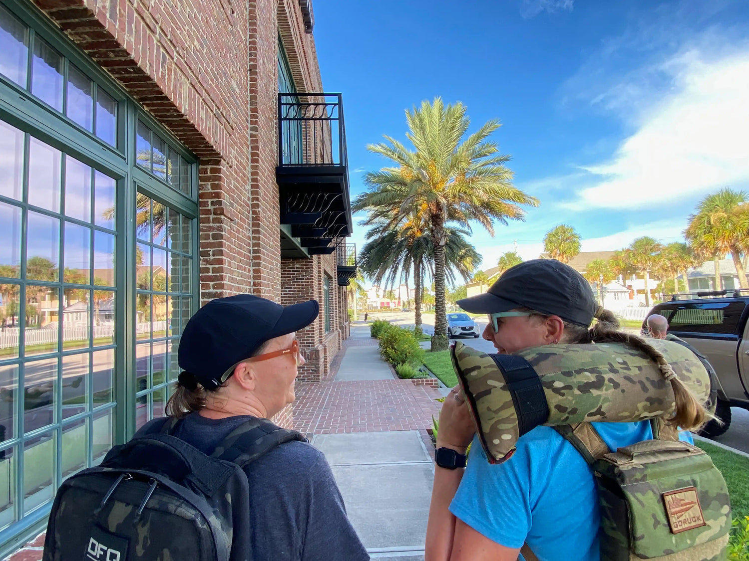 Two people rucking with GORUCK backpacks and sandbag, walking by brick building and palm trees