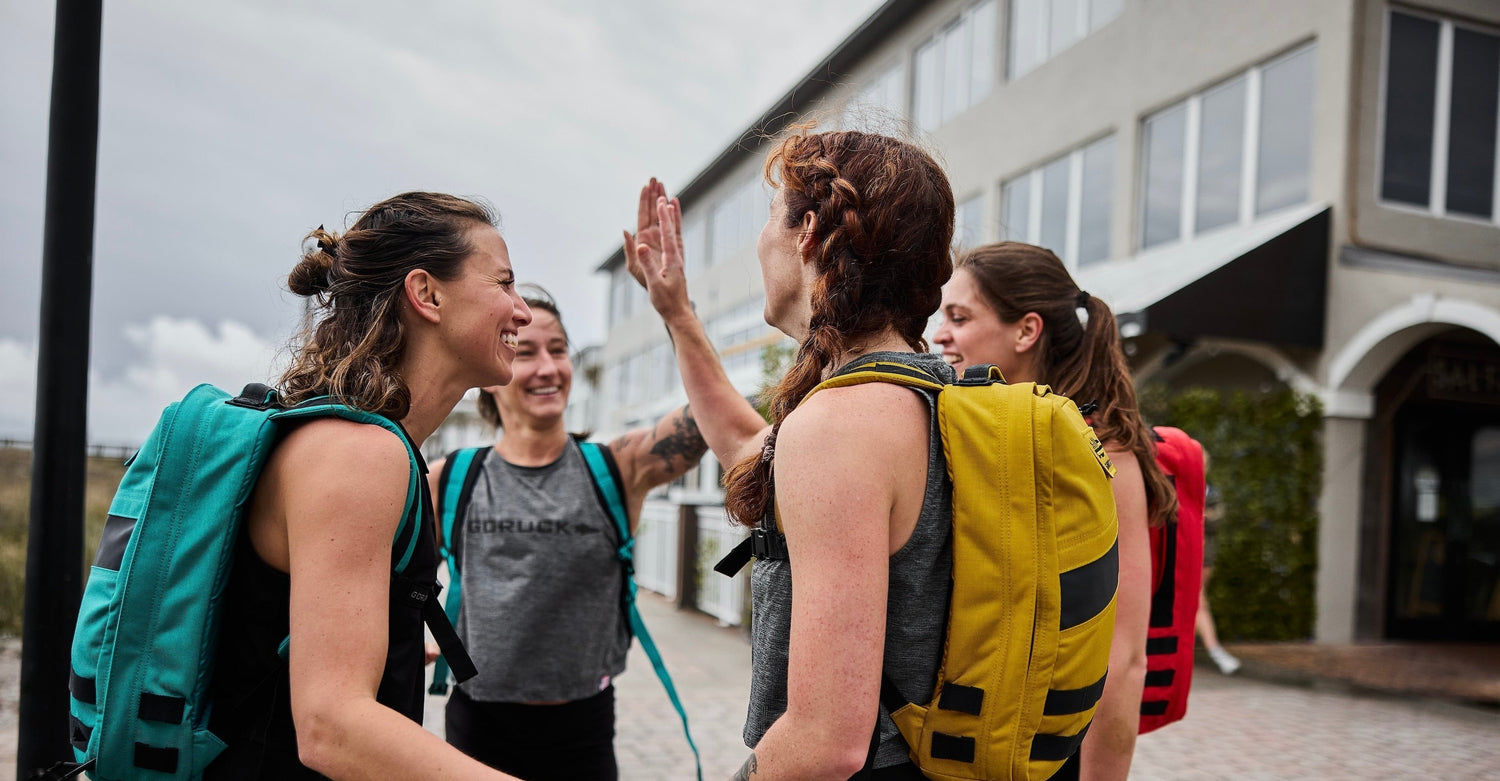 Group of women outdoors wearing colorful GORUCK backpacks celebrating with high fives near a building