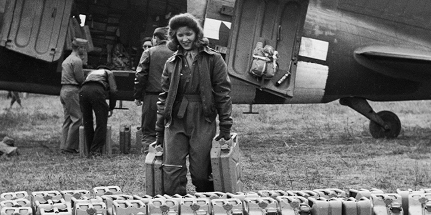 Vintage black and white photo of a woman carrying two jerry cans near a military airplane