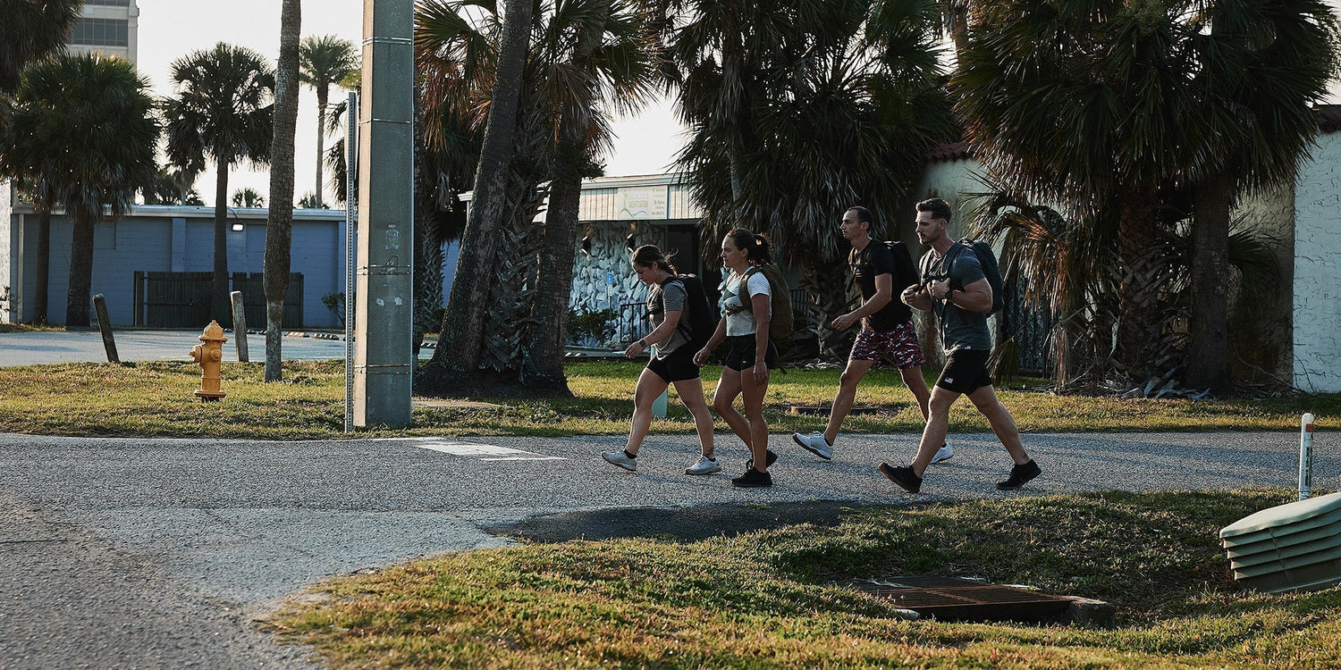 Group of people rucking with GORUCK backpacks, walking outdoors by palm trees and buildings