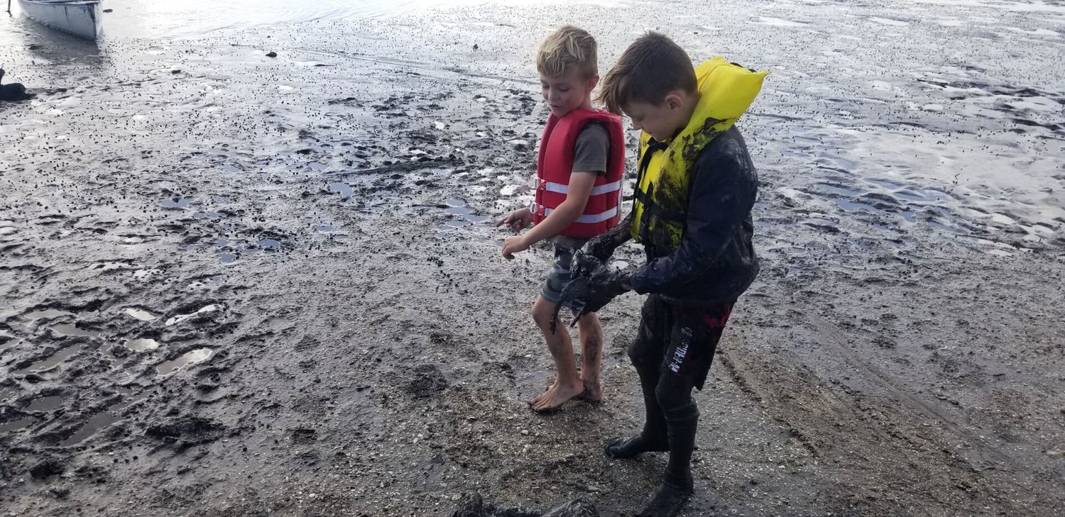 Two boys in life jackets playing barefoot in muddy lakeshore mud on a cloudy day