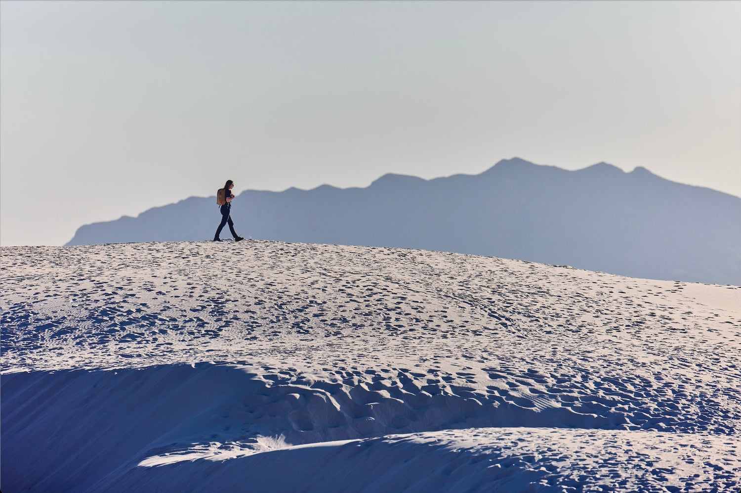 Person hiking alone on sandy dunes with mountainous background in soft light