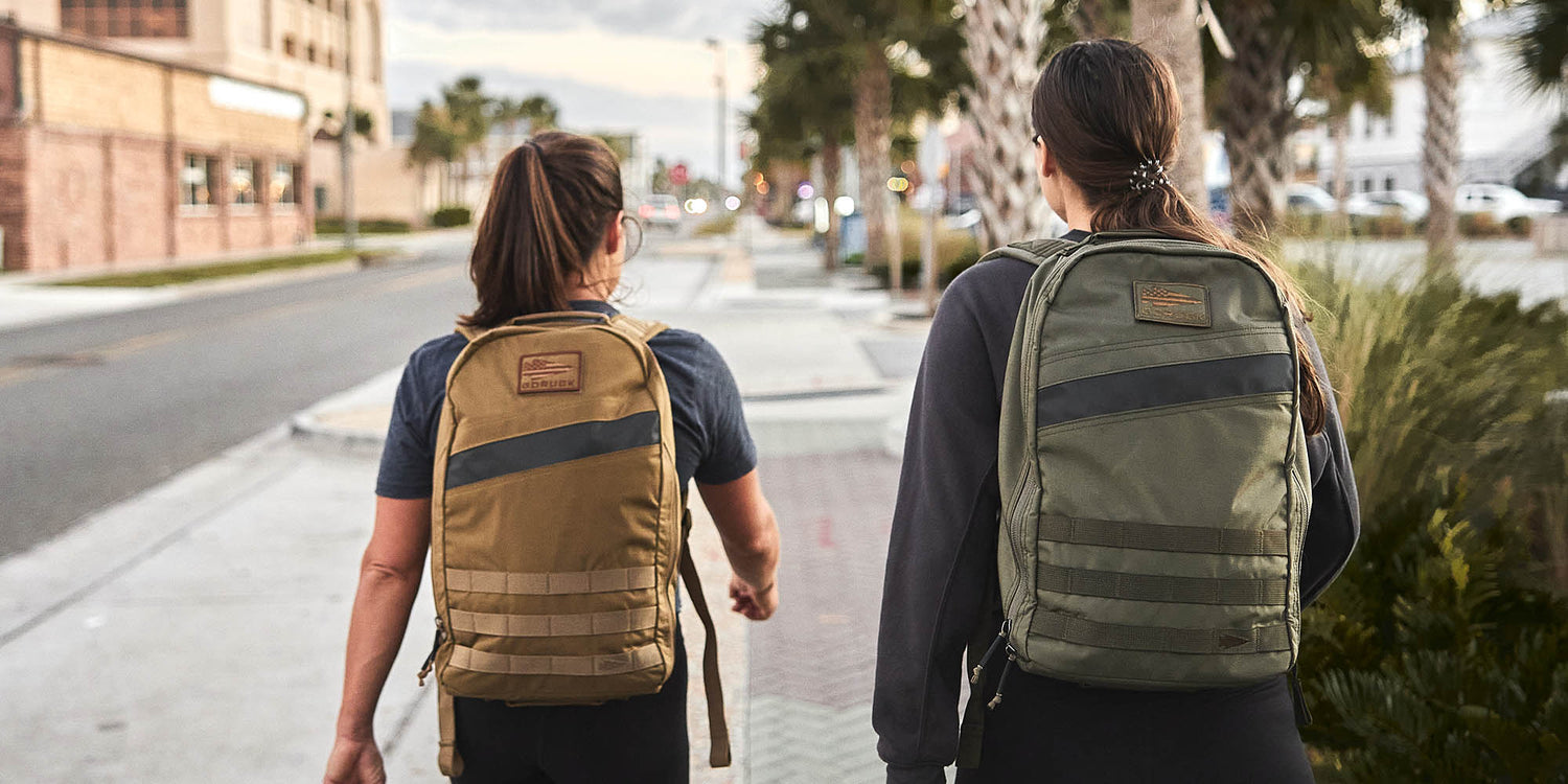 Two women rucking with GORUCK backpacks on a city sidewalk surrounded by palm trees