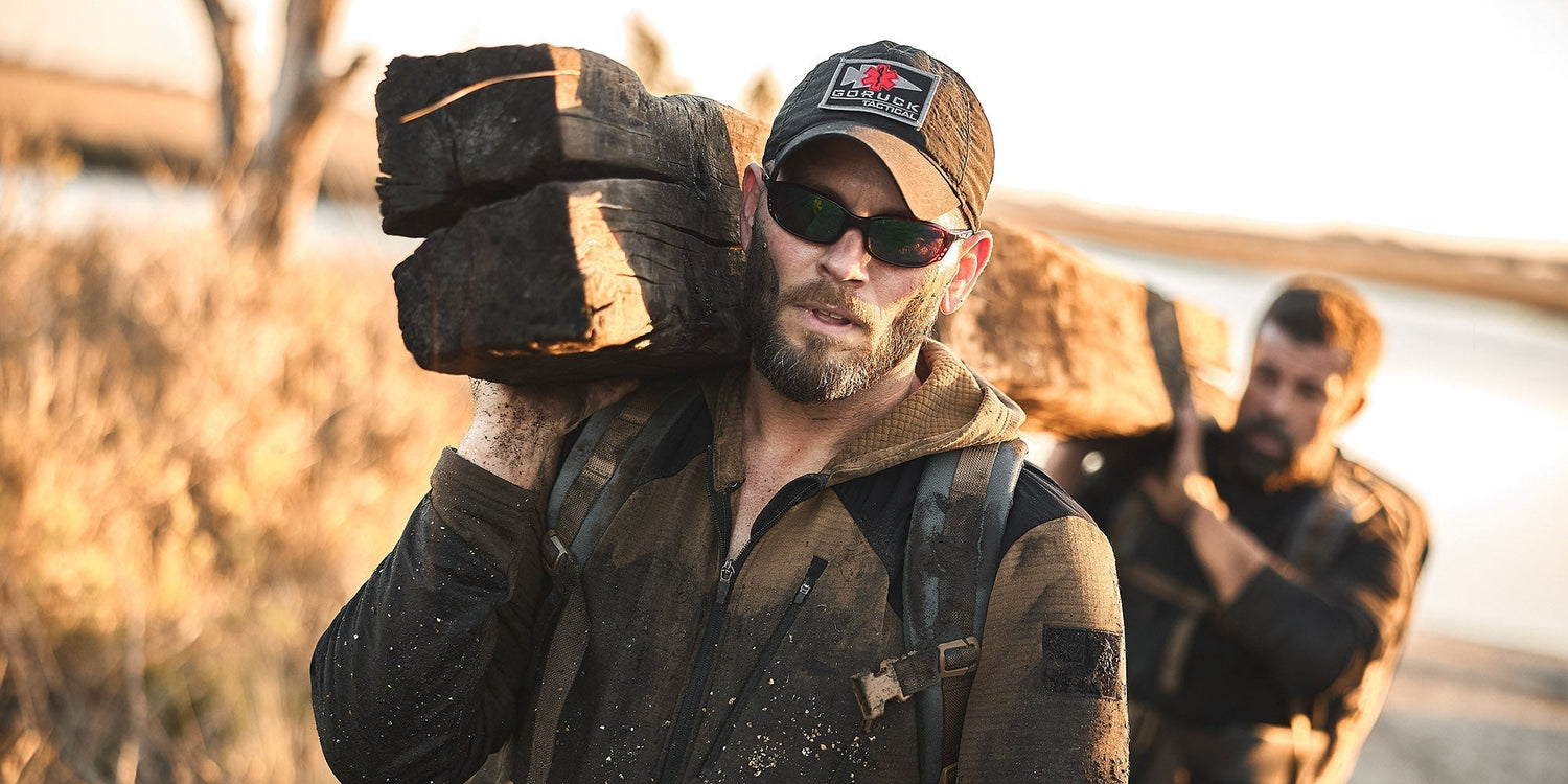 GORUCK team carrying heavy logs outdoors, wearing rugged rucking gear and tactical backpacks