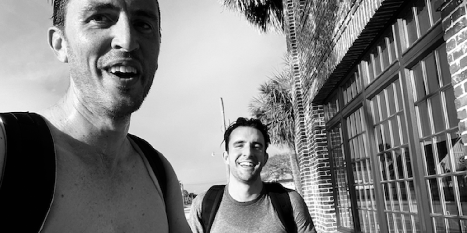 Two men with GORUCK backpacks smiling outdoors near a brick building, rucking together