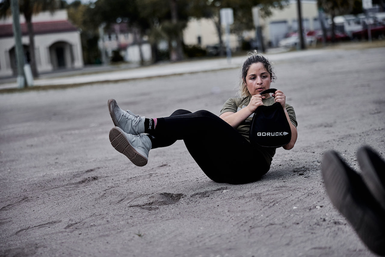 Woman performing outdoor Russian twist exercise with GORUCK kettlebell on dirt ground
