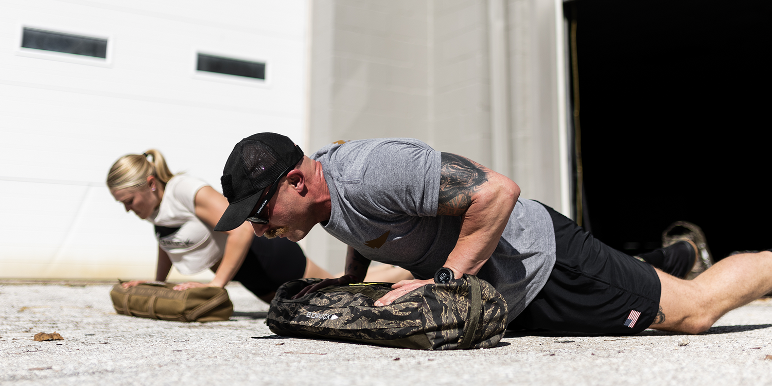 Man and woman doing push-ups outdoors with sandbag workout gear on concrete