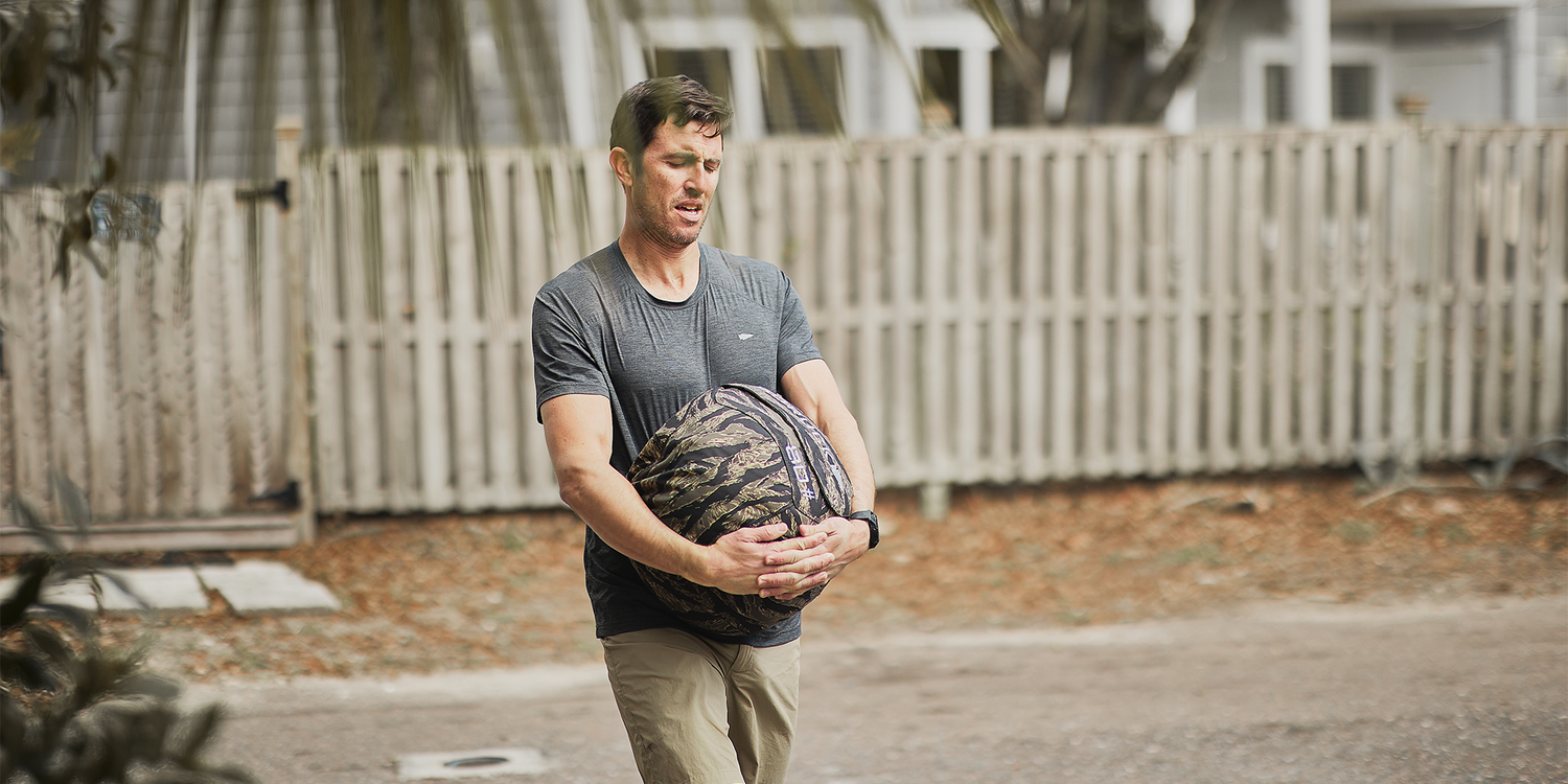 Man outdoors holding a heavy camouflage sandbag, wearing gray shirt and beige pants