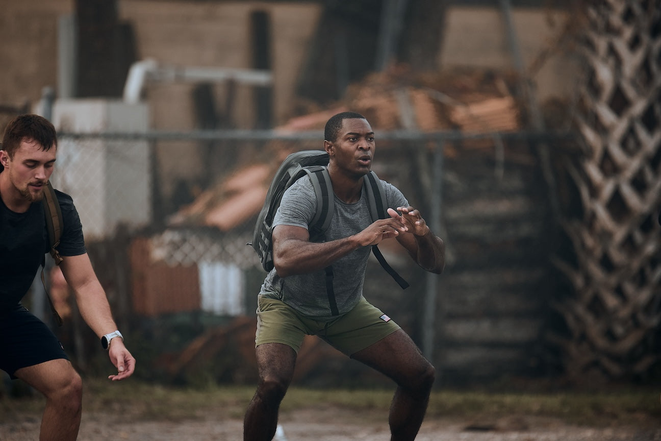 Two men outdoors doing squats with GORUCK rucksacks, showing tough rucking gear in action