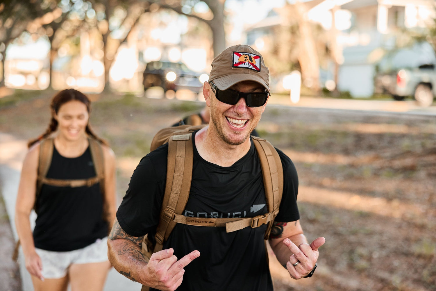Man smiling outdoors wearing GORUCK gear and backpack, woman rucking behind on sunny day