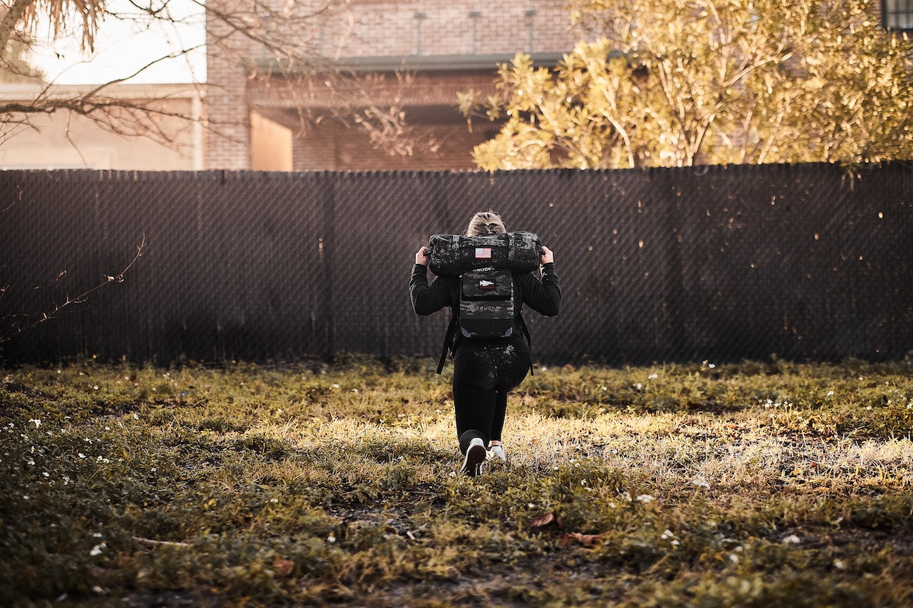 Person rucking outdoors with GORUCK backpack and sandbag, training in grassy field