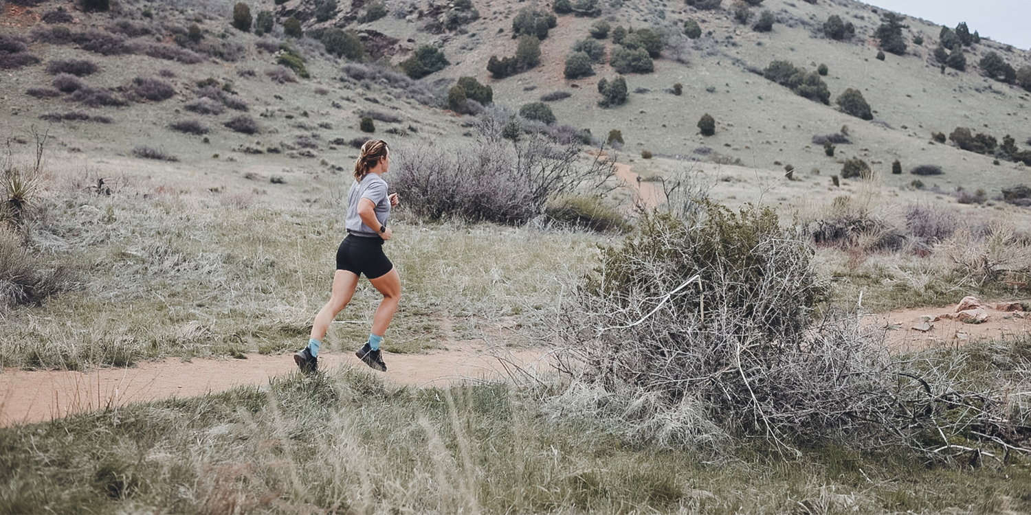 Woman trail running on dirt path in dry grassy mountain landscape wearing black shorts and gray shirt