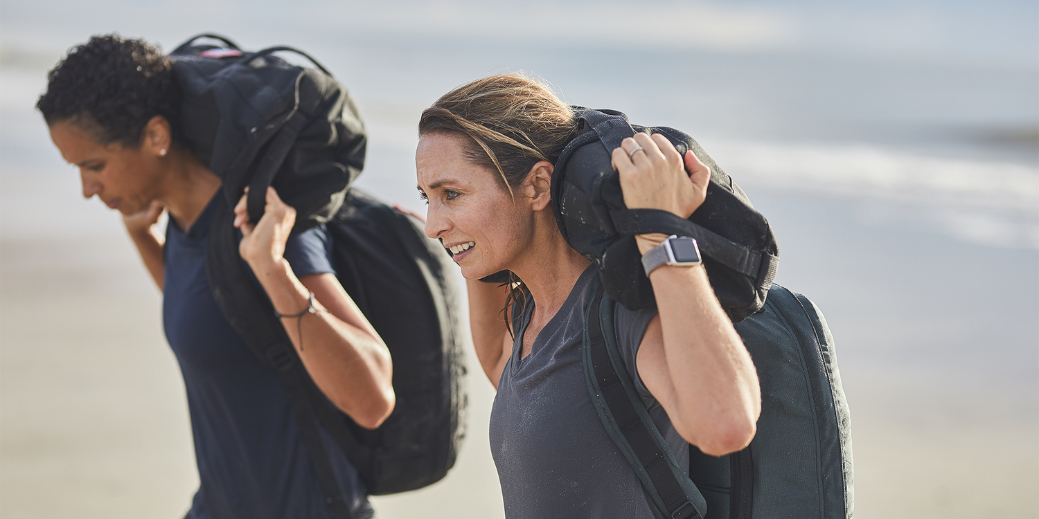 Two women rucking with GORUCK backpacks on a beach, focused and active outdoors
