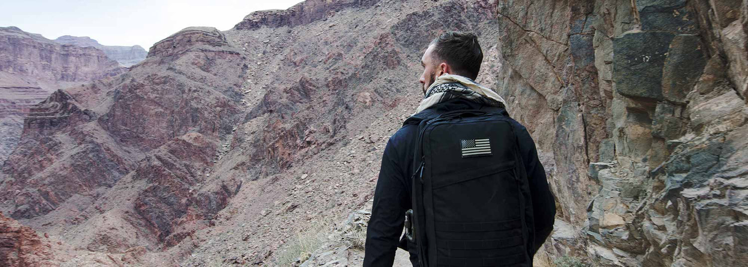 Man wearing black GORUCK backpack with American flag patch hiking in rugged desert canyon terrain