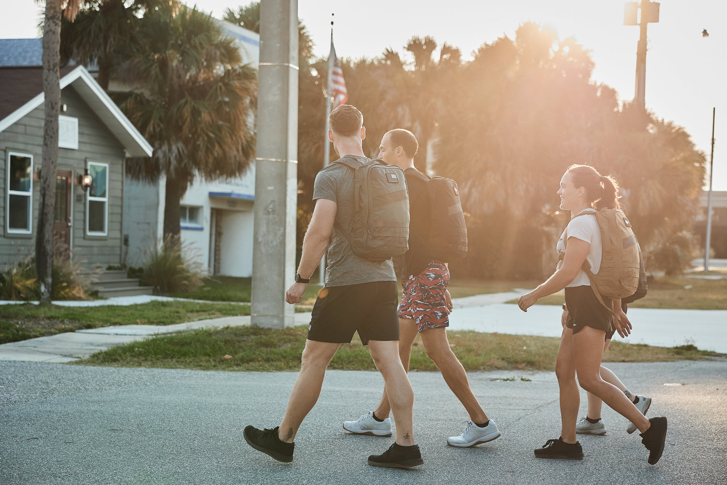 Four young adults with backpacks walk together on a sunny street near houses and palm trees.