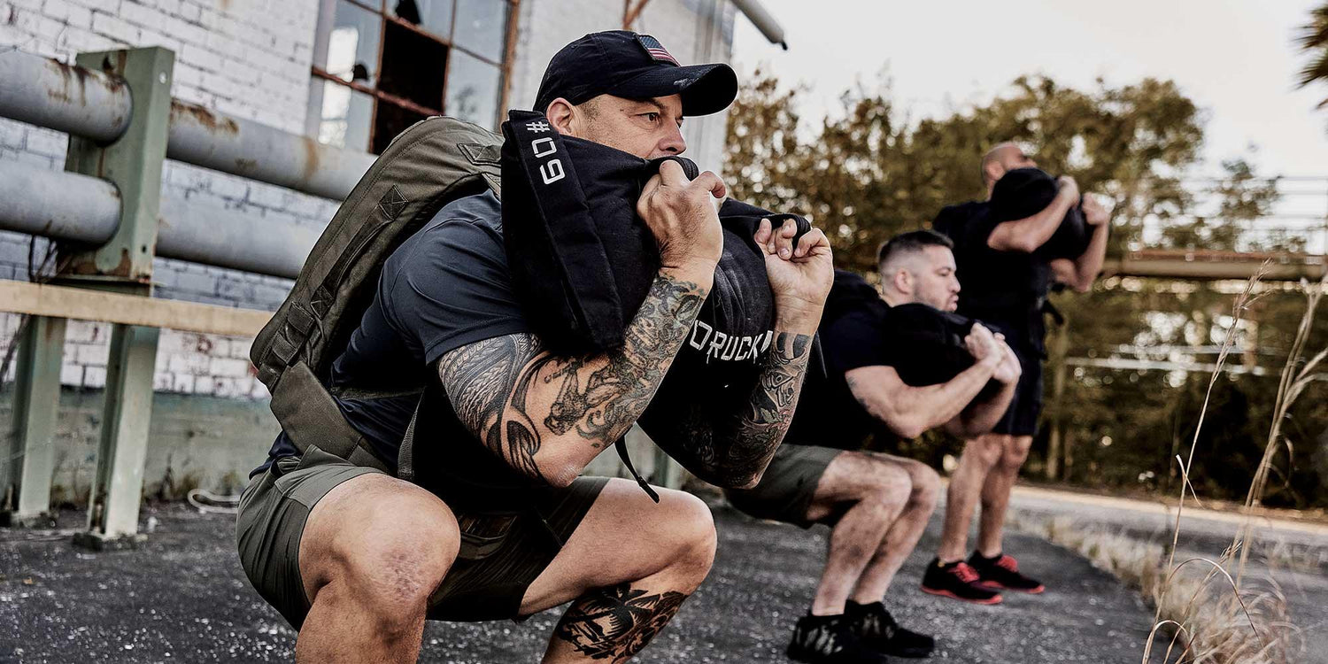Group of men outdoors doing squats with GORUCK sandbags, showcasing rugged fitness training gear.