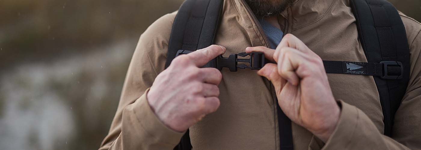 Person securing black GORUCK backpack chest strap outdoors, wearing brown jacket, close-up view