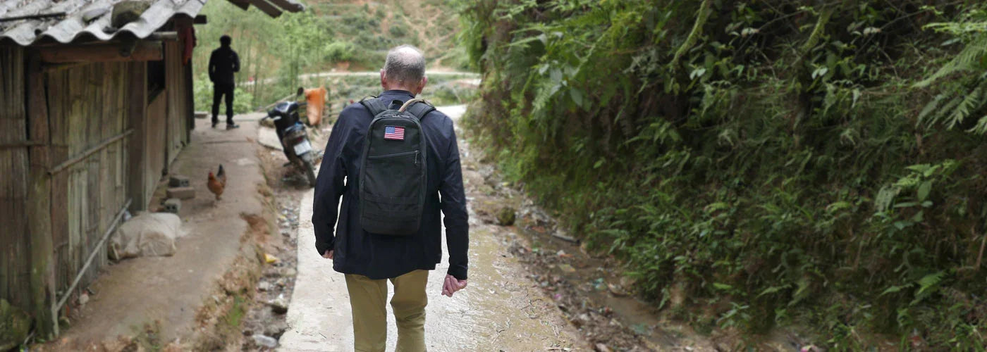 Man wearing black GORUCK backpack with American flag patch hiking on rural dirt path beside wooden house and green foliage