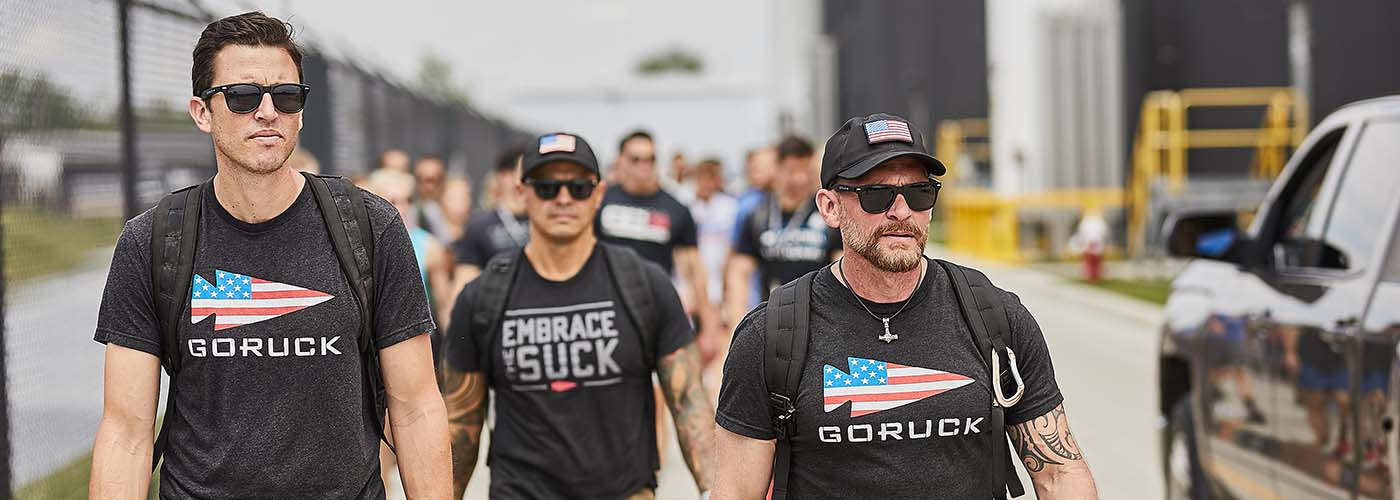 Three men wearing sunglasses and GORUCK shirts walk together outdoors among a group of people.