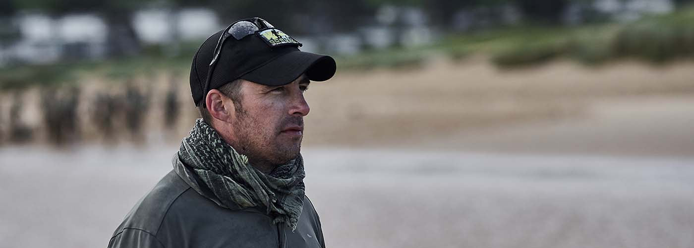 Man in outdoor gear and a cap stands on a sandy beach, looking into the distance.