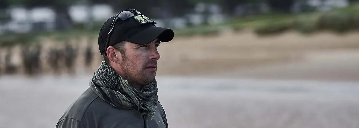 Man wearing GORUCK black cap, sunglasses, and camouflage scarf outdoors on sandy beach