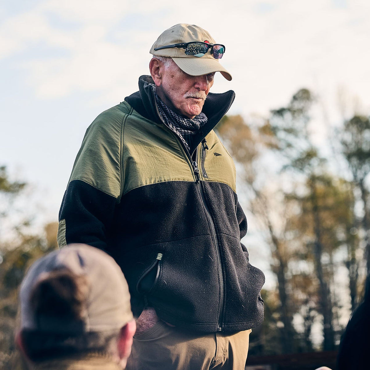 An older man wearing the Men’s Task Force Dagger Jacket - Polartec Fleece and sunglasses stands outdoors, addressing a group with trees behind him.