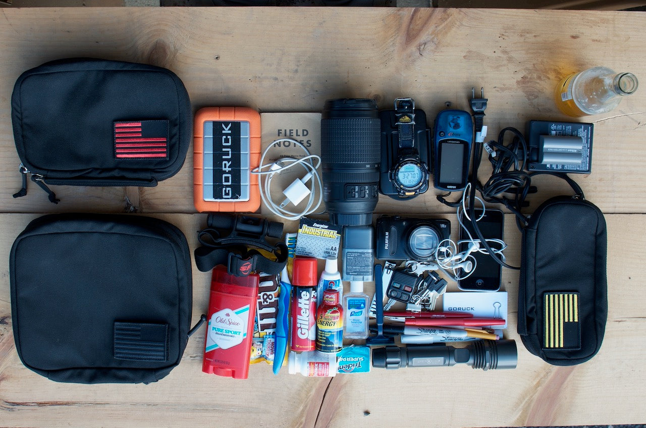 Assorted travel essentials, gadgets, and toiletries organized on a wooden surface with three black pouches.