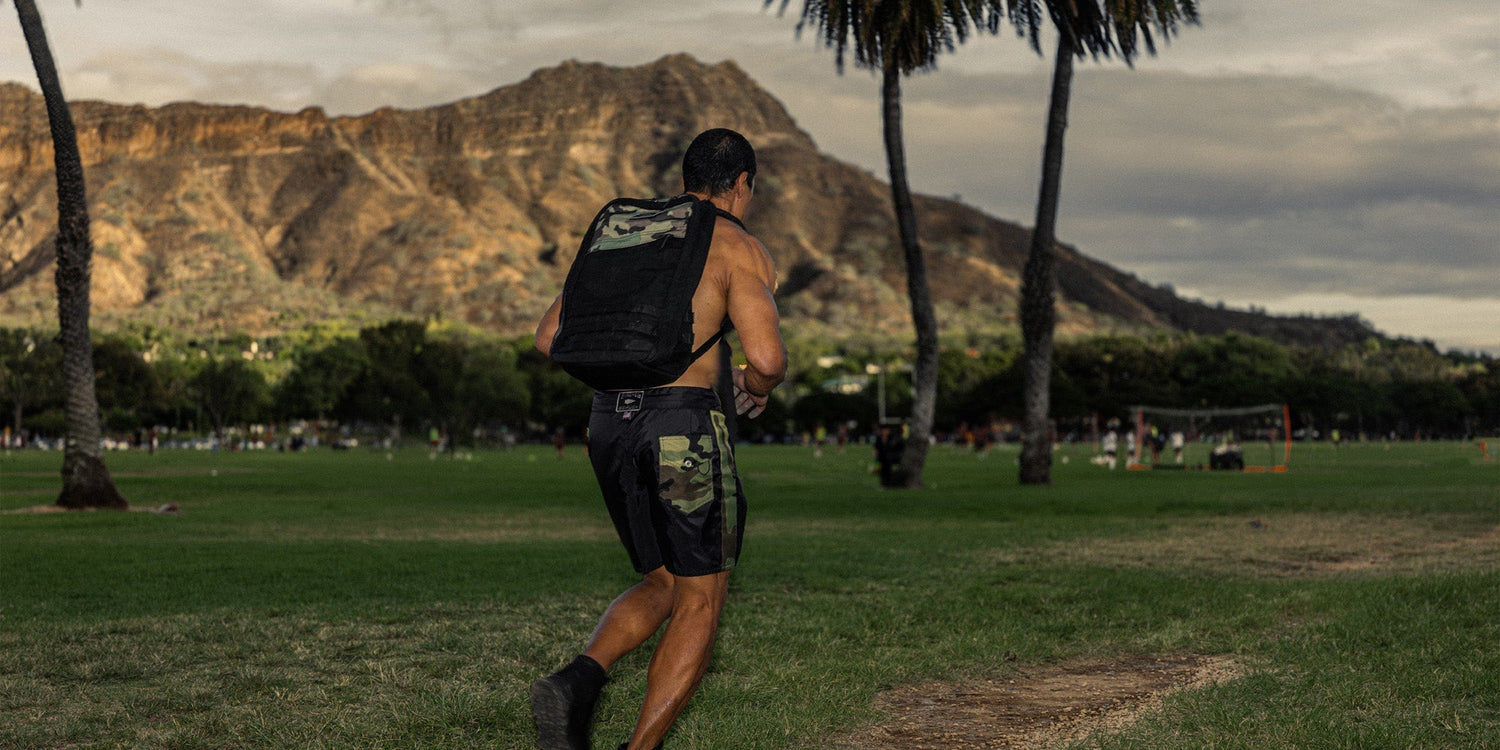 Athletic man in camo shorts carrying a GORUCK backpack in a grassy park with mountain and palm trees background
