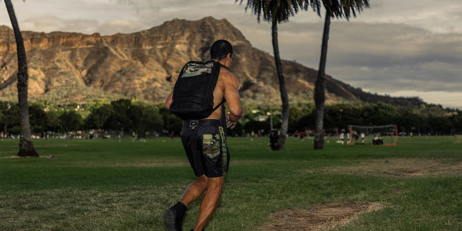 Man rucking with GORUCK backpack and camo shorts on a grassy field with mountains in background