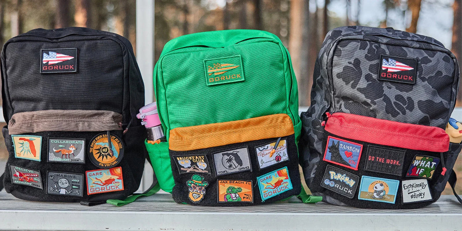 Three GORUCK rucking backpacks in black, green, and camo with colorful event patches attached
