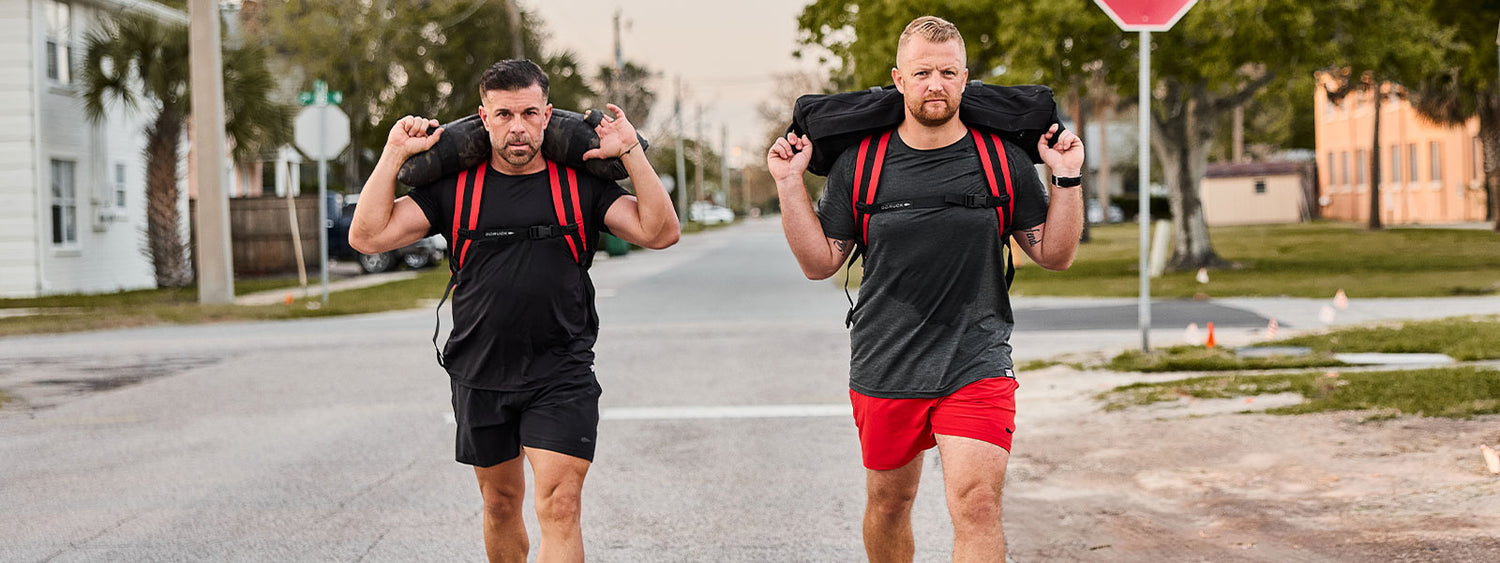 Two men in athletic clothing carry weighted bags on their shoulders while walking on a street.