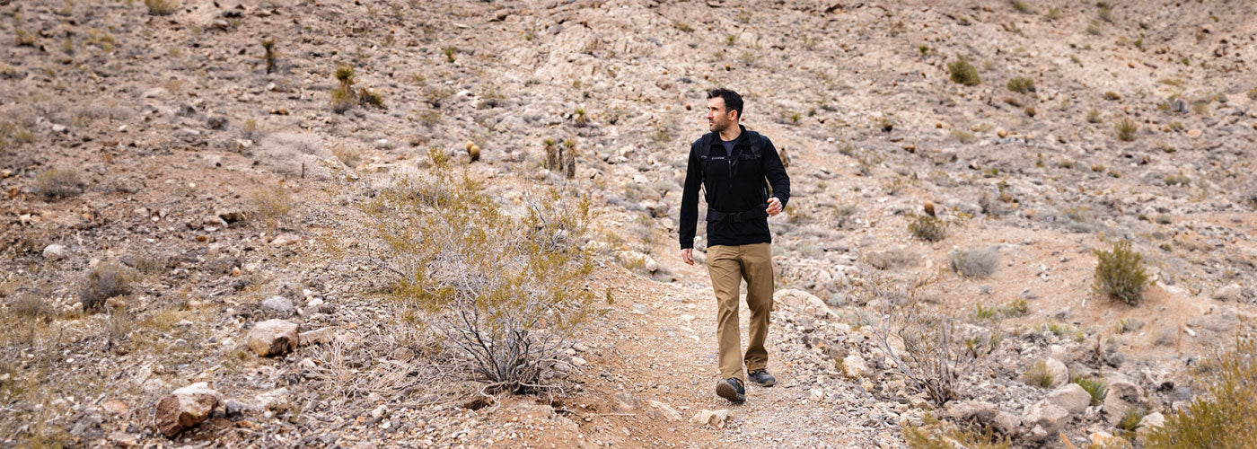 Man wearing GORUCK rugged gear walking on rocky desert trail with dry bushes