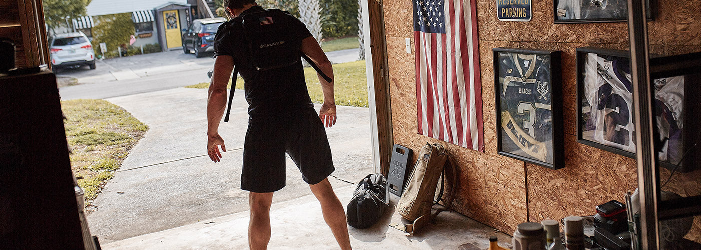 Man wearing GORUCK backpack training with weights in a garage decorated with American flag and sports memorabilia