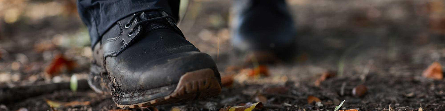 Close-up of black tactical boots walking on a dirt trail with fallen leaves, outdoors rugged footwear
