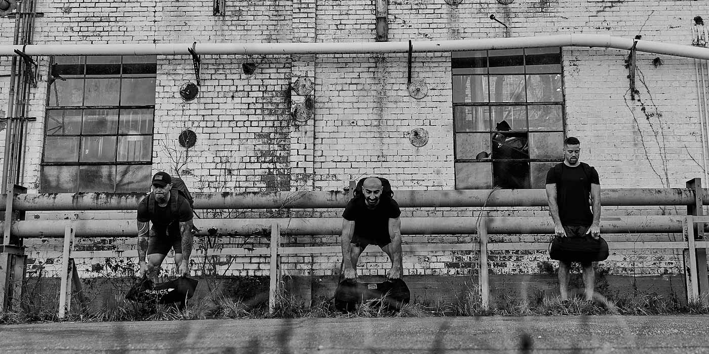 Three men with GORUCK bags performing outdoor strength training against a weathered brick wall backdrop