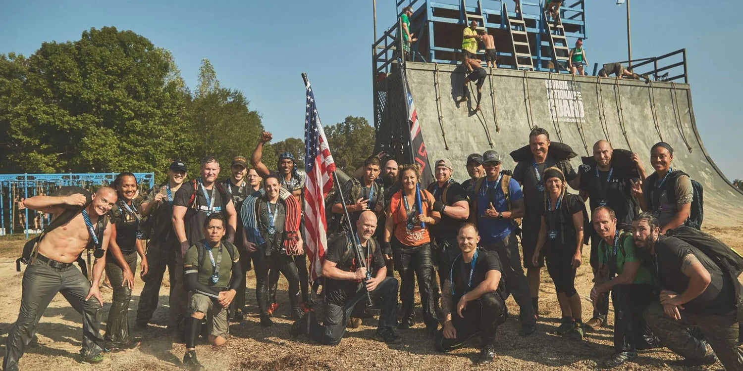 GORUCK rucking team posing with American flag after outdoor obstacle event, muddy and smiling